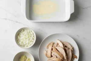 Zutaten für Puten-Auflauf: Gewürfelte Zwiebeln, Knoblauch und Putenstreifen neben einer eingefetteten weißen Keramik-Auflaufform.